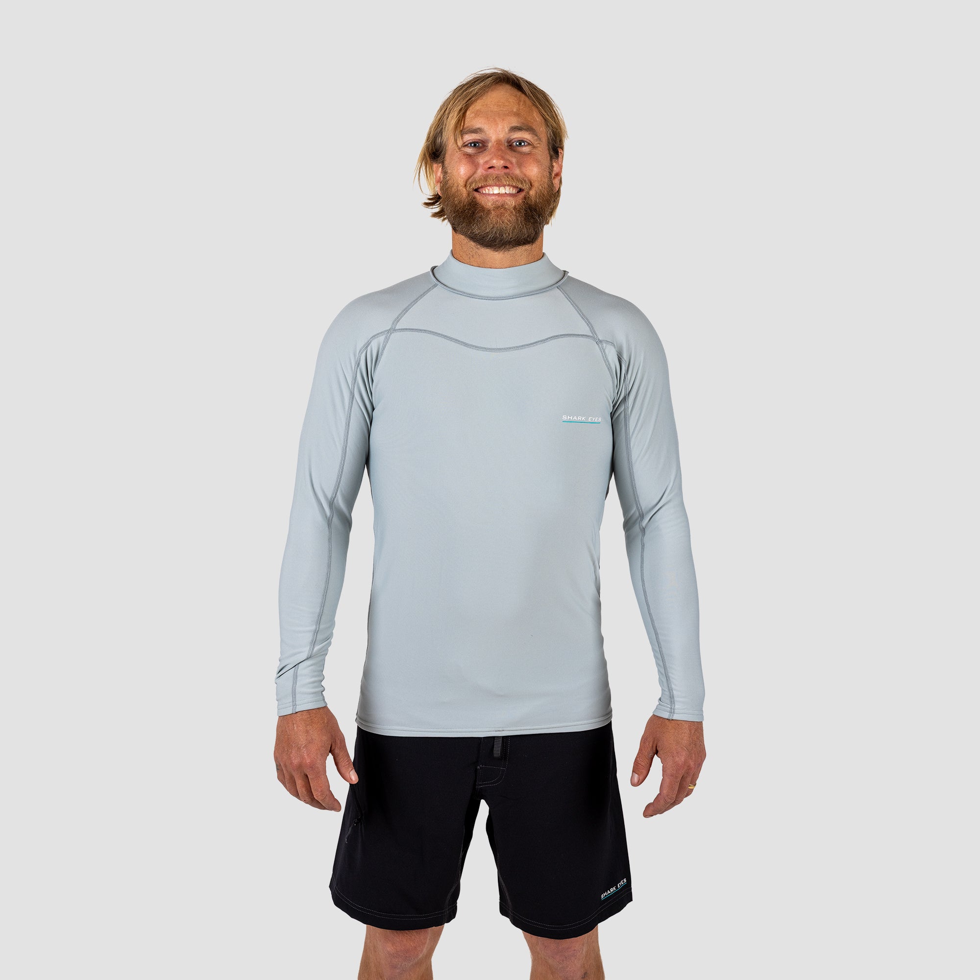 Man wearing a light gray long-sleeve Rash vest and black shark eyes instinct boardshorts on a light grey background