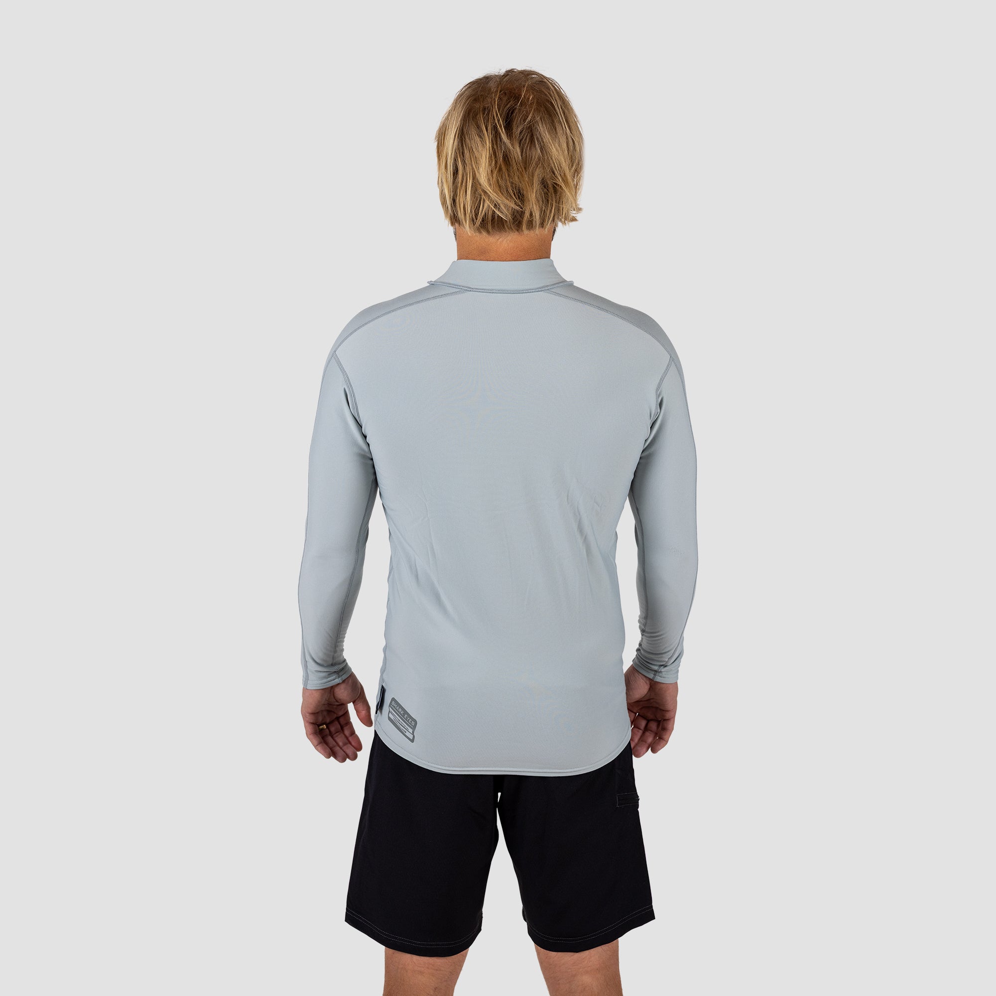 Man wearing a light gray long-sleeve Rash vest and black shark eyes instinct boardshorts on a light grey background