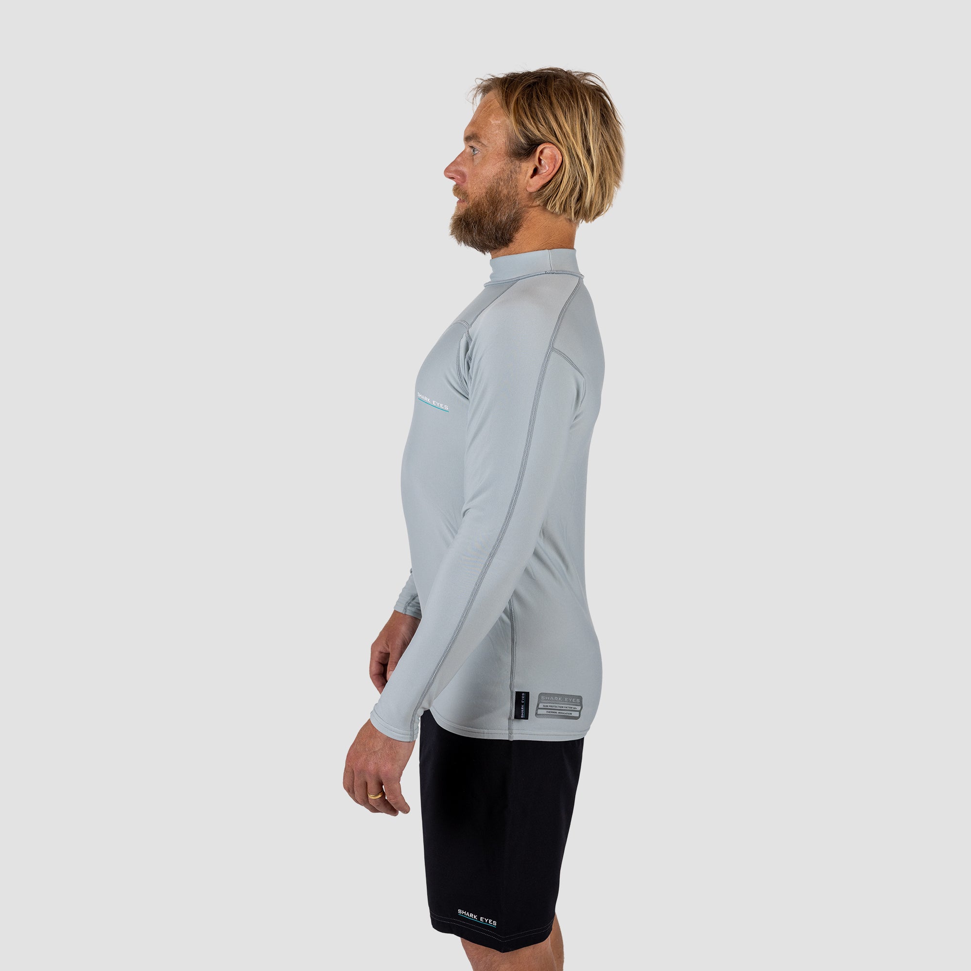 Man wearing a light gray long-sleeve Rash vest and black shark eyes instinct boardshorts on a light grey background